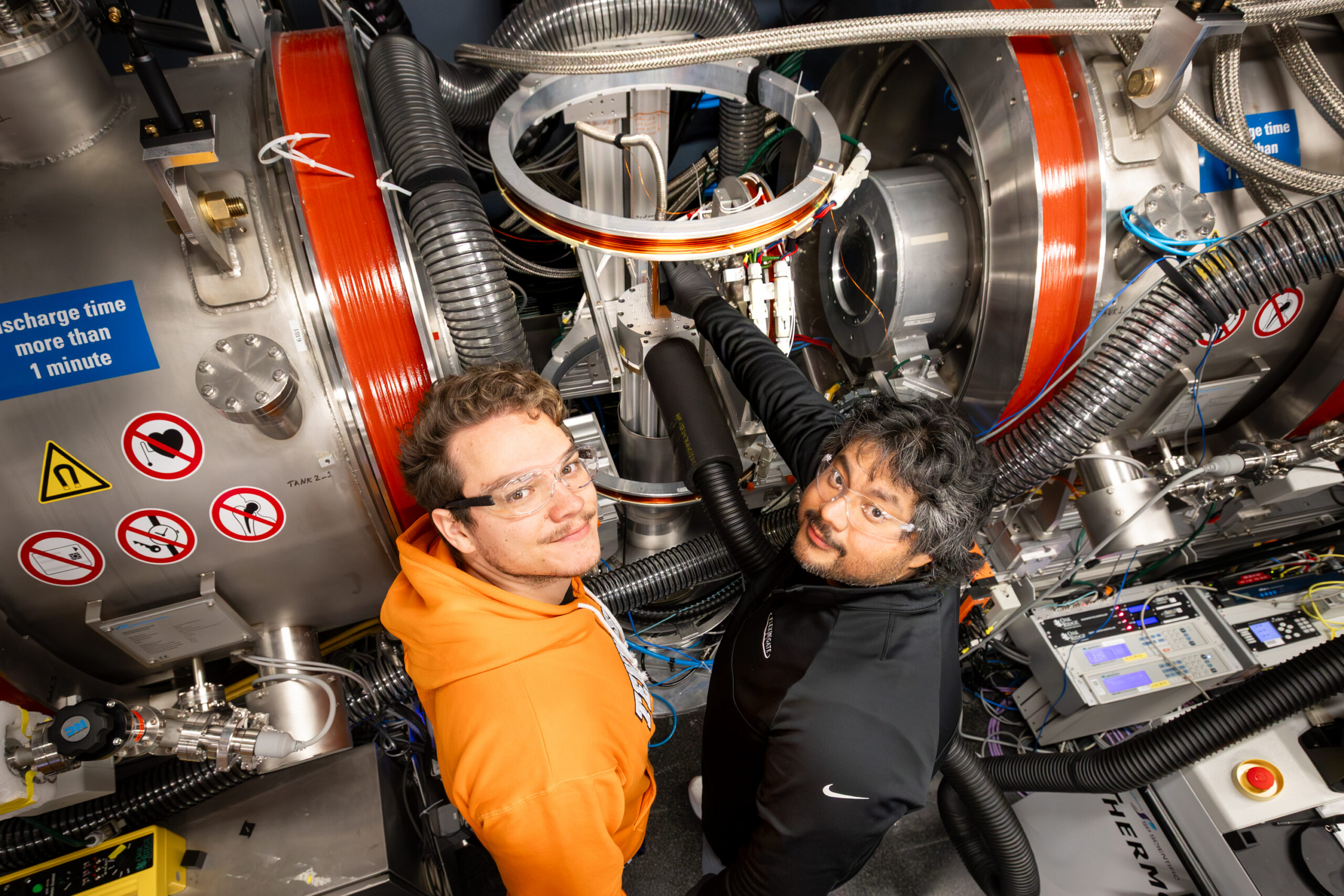 Two University of Windsor doctoral students, Stuart Castillo and Maks Dziura, wearing safety glasses, stand in front of the Neutron Spin Echo instrument at the Spallation Neutron Source, surrounded by coils, cables, and stainless-steel experimental equipment.